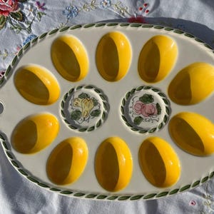 May include: An oval ceramic egg serving platter with a cream-colored base and a green vine-patterned border. The platter features ten bright yellow, oval-shaped indentations for eggs and two central circular decorative motifs with fruit illustrations.