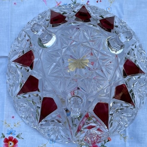Puede incluir: Un cuenco redondo de cristal transparente con un patrón geométrico facetado en forma de estrella y acentos triangulares de color rojo intenso integrados en el diseño. El cuenco se apoya sobre tres pequeñas patas de cristal transparente.