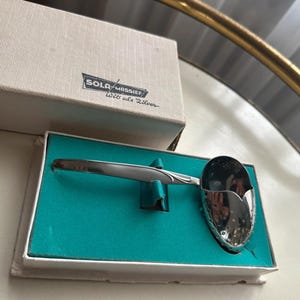 Op de afbeelding: Een zilverkleurige theezeef van metaal met een geperforeerde kom en een glad handvat, gepresenteerd in een witte rechthoekige doos met een fel turquoise inzetstuk. Het deksel bevat de tekst SOLA MASSIEF en Wits als Zilver in zwarte opdruk.