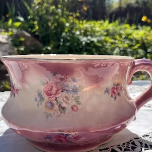 Op de afbeelding: Een keramische po met een glanzende roze verloopafwerking en een centraal bloemenboeketmotief. De pot heeft een brede, uitlopende rand, een gebogen handvat aan de rechterkant en kleine bloemaccenten verspreid over het buitenoppervlak.