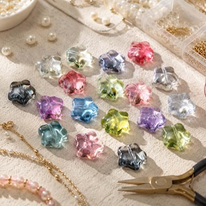 Può includere: Una collezione di perline traslucide a forma di stella in vari colori, tra cui rosa, blu, verde, viola e trasparente. Le perline sono disposte su una superficie ruvida insieme a una catenina dorata, perle bianche sfuse e una pinza.