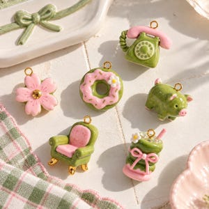 May include: A collection of six small charms featuring a pink flower, a rotary telephone, a donut, a pig, an armchair, and a drink, each with a gold-tone metal loop for attachment. The charms are crafted in shades of sage green and pastel pink.