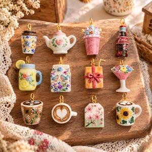 May include: A collection of twelve miniature ceramic charms featuring various designs including a teapot, a soda bottle with the text Loda, a milk carton, a gift box, a martini glass with flowers, and several mugs with floral and heart patterns.
