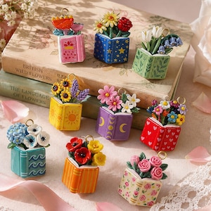 Puede incluir: Una colección de nueve pequeños dijes, cada uno con un libro abierto en miniatura con un ramo de flores coloridas que emerge de las páginas. Los dijes muestran varios patrones y colores, incluyendo diseños florales, celestiales y geométricos.