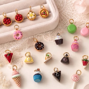 Könnte beinhalten: Eine Sammlung von Miniatur-Anhängern im Lebensmittel-Design, die verschiedene Desserts wie Obsttörtchen, Donuts, Cupcakes, Kuchenstücke, Eiswaffeln, Macarons und einen Lutscher darstellen. Jeder Anhänger hat einen goldfarbenen Metallring zur Befestigung.