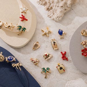 May include: A collection of gold-tone metal charms featuring various shapes including palm trees, starfish, seashells, fish, flowers, and playing cards, some with enamel accents, displayed alongside three pairs of flip-flops in cream, gray, and navy blue.