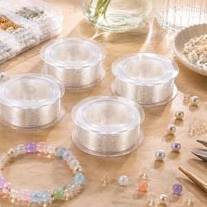 Puede incluir: Tres carretes de plástico transparente con cordón elástico fino para bisutería, dispuestos junto a una pulsera de cuentas terminada, una caja organizadora de plástico con cuentas variadas, un cuenco pequeño con cuentas sueltas y alicates de metal.