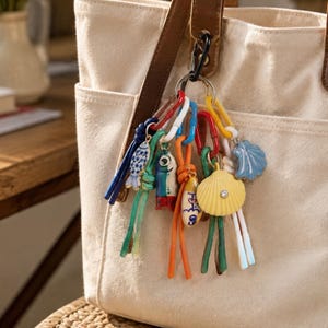 Può includere: Una borsa tote in tela beige con manici in pelle marrone presenta un portachiavi decorativo. Il portachiavi è composto da un moschettone nero che sostiene maglie in plastica colorate, cordini annodati blu, verdi e arancioni e ciondoli a forma di pesci e conchiglie.