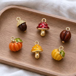 Pode incluir: Um conjunto de seis pequenos pingentes de metal com designs de temática de outono, incluindo uma bolota castanha, um cogumelo vermelho com pontos brancos, uma noz castanha, uma abóbora laranja com folha verde, um cogumelo amarelo e uma abóbora.