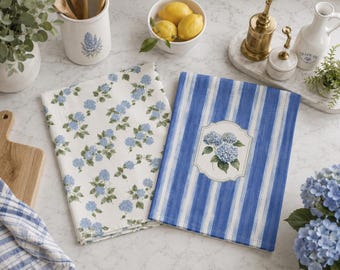 Puede incluir: Dos paños de cocina rectangulares. Uno presenta un fondo blanco roto con un patrón repetitivo de flores de hortensia azules y hojas verdes. El segundo tiene rayas verticales azules y blancas con una etiqueta decorativa central.