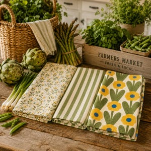 Puede incluir: Tres pa&ntilde;os de cocina rectangulares plegados con patrones distintos: uno con peque&ntilde;as flores amarillas y azules, otro con rayas verticales verdes y blancas, y otro con un motivo de flores amarillas grandes y hojas verdes sobre fondo crema.