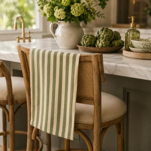 Puede incluir: Un pa&ntilde;o de cocina con rayas verticales de color verde salvia y crema colgado sobre el respaldo de un taburete de madera con respaldo de rejilla y asiento tapizado en beige, situado en una cocina con encimera de m&aacute;rmol.