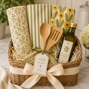 Puede incluir: Una cesta de mimbre que contiene tres pa&ntilde;os de cocina plegados con patrones florales y de rayas, una cuchara de madera, una esp&aacute;tula de madera ranurada y una botella de 500 ml de aceite de oliva virgen extra. Una etiqueta dice A Gift for you MADE WITH LOVE.