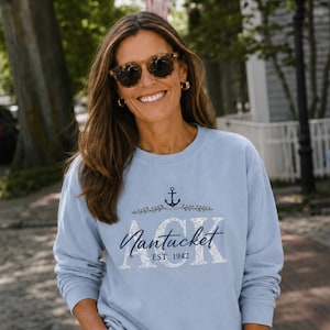 May include: A light blue crewneck sweatshirt featuring a dark blue anchor icon above the text ACK, with the word Nantucket written in cursive script across the center and the text EST. 1942 printed below in a smaller, clean sans-serif font.