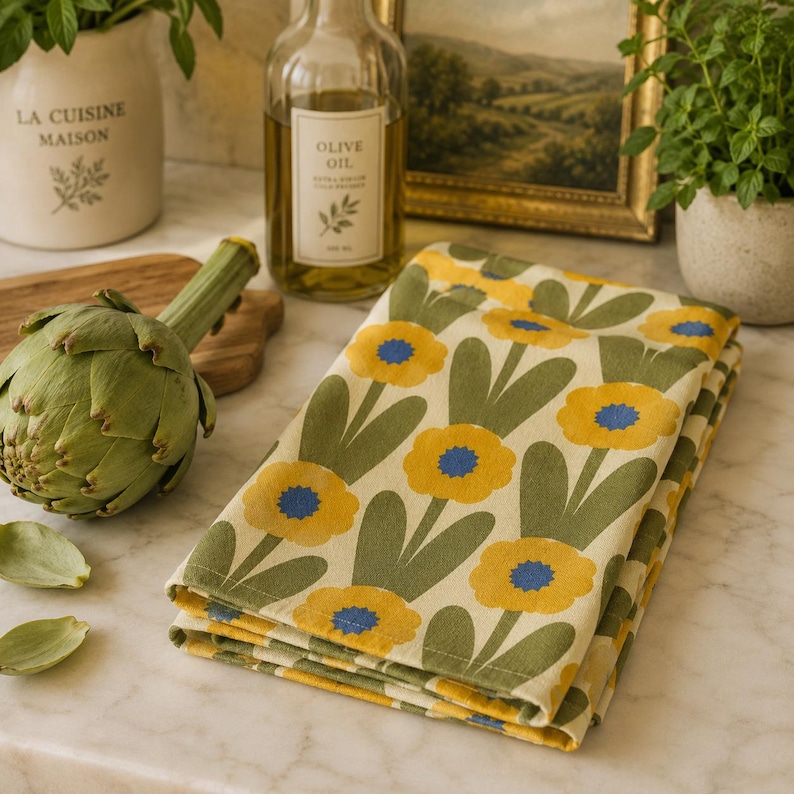 Puede incluir: Un pa&ntilde;o de cocina plegado con un patr&oacute;n repetitivo de flores amarillas con centros azules y tallos verdes sobre un fondo crema. La tela tiene un tejido visible y est&aacute; colocada junto a una alcachofa verde entera y una botella de aceite de oliva.