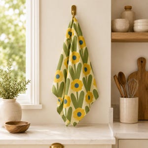 Puede incluir: Un pa&ntilde;o de cocina con un patr&oacute;n repetitivo de flores amarillas con centros azules y tallos verdes sobre un fondo crema. El pa&ntilde;o cuelga de un gancho de lat&oacute;n en la pared, mostrando un dise&ntilde;o floral vibrante con l&iacute;neas y formas claras.