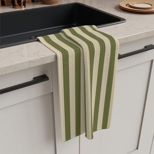 Puede incluir: Un pa&ntilde;o de cocina rectangular con un patr&oacute;n de rayas verticales en tonos alternos de verde oliva y crema. La tela tiene un tejido texturizado tipo gofre y est&aacute; colgado sobre el borde de una encimera de cocina junto a un fregadero negro.