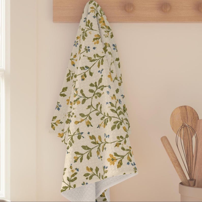 Puede incluir: Un pa&ntilde;o de cocina de color crema con un patr&oacute;n repetitivo de enredaderas verdes, peque&ntilde;as flores amarillas y diminutas flores azules. El pa&ntilde;o cuelga verticalmente de un soporte de madera, mostrando una textura suave y un dise&ntilde;o bot&aacute;nico.