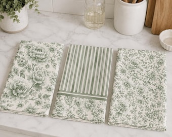 Puede incluir: Tres paños de cocina rectangulares con patrones en verde salvia sobre fondo crema. Uno muestra flores grandes, otro rayas verticales sobre un borde floral, y el tercero presenta un estampado floral denso de pequeña escala.