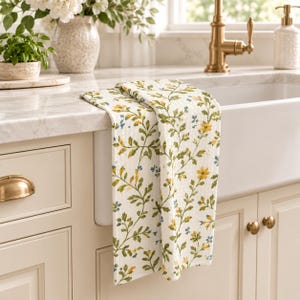 Puede incluir: Un pa&ntilde;o de cocina blanco con un patr&oacute;n bot&aacute;nico repetitivo de tallos verdes, flores amarillas y peque&ntilde;as flores azules. El pa&ntilde;o est&aacute; colgado sobre el borde de un fregadero blanco, mostrando una tela texturizada con un estampado floral.