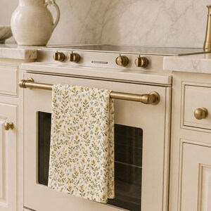 Puede incluir: Un pa&ntilde;o de cocina rectangular con un patr&oacute;n repetitivo de peque&ntilde;as flores amarillas, hojas verdes y diminutas flores azules sobre un fondo de color crema, colgado del asa horizontal de lat&oacute;n de un horno de cocina color crema.