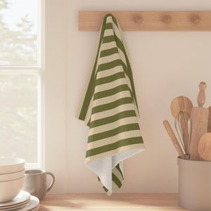 Puede incluir: Un pa&ntilde;o de cocina rectangular con un patr&oacute;n repetitivo de rayas horizontales en color verde oliva y crema. La tela presenta un tejido texturizado y est&aacute; colgada de un perchero de madera, mostrando un dise&ntilde;o de rayas uniforme en toda la pieza.