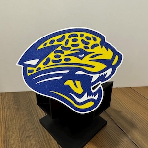 May include: A glittery cutout of a jaguar head logo in blue and yellow, featuring a stylized profile with an open mouth and spotted pattern. The cutout is mounted on a black, multi-tiered, geometric stand with a square base and vertical dividers.