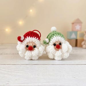 Könnte beinhalten: Zwei kleine, runde, gehäkelte Ornamente in Form von Weihnachtsmann-Gesichtern. Eine Figur trägt eine rote Mütze mit weißem Bommel, die andere eine grüne Mütze mit weißem Bommel. Beide haben gehäkelte weiße Bärte, kleine schwarze Augen und rote Nasen.