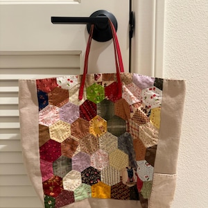 Op de afbeelding: Een tote bag met een patchworkontwerp van veelkleurige stoffen zeshoeken in diverse patronen, waaronder bloemen-, kersen- en ruitjesprints. De tas heeft effen beige zijpanelen en twee rode lederen handvatten aan de bovenrand.