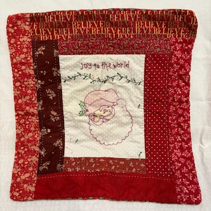 May include: A square quilted pillow cover featuring a central white panel with an embroidered Santa Claus face and the text "Joy to the world". The border consists of various red fabric strips with floral, polka dot, and repeating "BELIEVE" patterns.