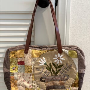 Könnte beinhalten: Eine rechteckige, gesteppte Tragetasche mit Patchwork-Design in Gelb-, Creme- und Weißtönen. In der Mitte befindet sich eine kreisförmige Applikation einer Topfblume mit weißen Blütenblättern und grünen Blättern. Braune Leder-Schulterriemen.
