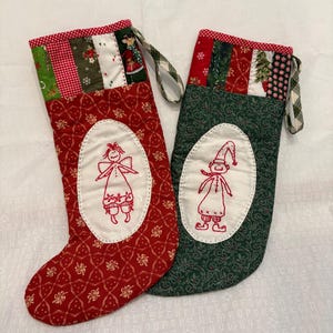 Könnte beinhalten: Zwei gesteppte Weihnachtsstrümpfe mit Aufhängeschlaufen. Der linke Strumpf ist rot mit einem weißen ovalen Aufnäher, der einen rot gestickten Engel zeigt. Der rechte Strumpf ist grün mit einem weißen ovalen Aufnäher, der einen rot gestickten Elfen zeigt.