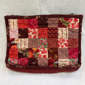 May include: A rectangular zippered pouch featuring a patchwork design composed of various fabric squares in shades of red, burgundy, and cream. The quilted textile displays a mix of floral, gingham, and solid patterns with a dark red border.