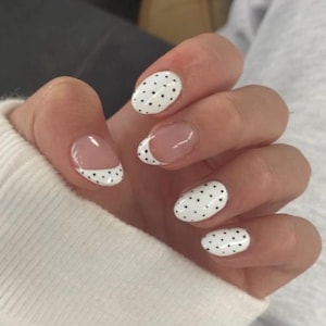 May include: Oval-shaped fingernails featuring a white base coat with small black polka dots. Some nails display a French manicure style with a clear nail bed and white tips covered in black dots, while others are fully painted white with black dots.