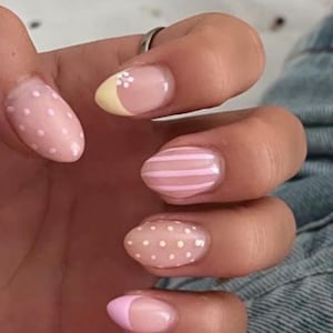 May include: Oval-shaped fingernails featuring a variety of designs including light pink with white polka dots, a pale yellow French tip with a small white flower, light pink and white vertical stripes, and a solid light pink French tip.