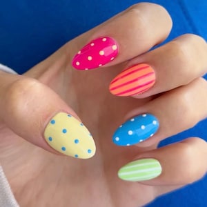 May include: Almond-shaped fingernails featuring a colorful manicure. The thumb is pale yellow with blue dots, the index finger is hot pink with yellow dots, the middle finger has orange stripes, the ring finger is blue with white dots, and the pinky is light green with white stripes.
