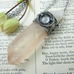 May include: A rose quartz crystal pendant necklace with a silver-toned chain. The crystal is a pale pink color and is faceted. The pendant is set in a decorative silver-toned metal setting. The necklace is displayed on a vintage book.