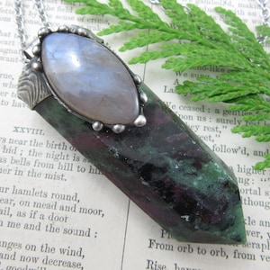 May include: A pendant necklace featuring a dark green and deep red zoisite crystal point topped with a smooth, oval-shaped iridescent moonstone cabochon set in a textured silver metal cap, suspended from a thin silver-toned metal chain.