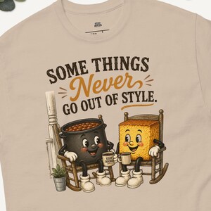 Pode incluir: Uma t-shirt bege de manga curta com um gráfico de uma panela de feijão e um quadrado de pão de milho sentados em cadeiras de balanço. O texto acima dos personagens diz: "SOME THINGS Never GO OUT OF STYLE." Gola redonda clássica.