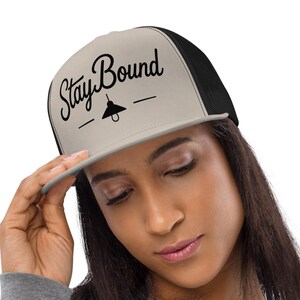May include: A beige and black trucker hat featuring a flat brim and a mesh back panel. The front panel displays the black text Stay Bound in a cursive script font, positioned above a small graphic of a hanging light bulb with horizontal lines.