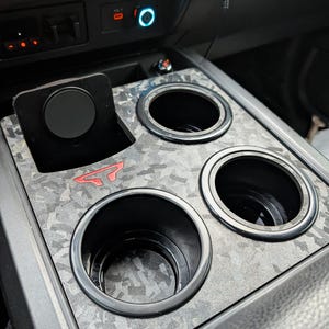 May include: A black vehicle center console insert featuring a gray and black marbled pattern with three circular cup holders and a rectangular slot. A small red geometric logo is embedded on the surface near the cup holders.