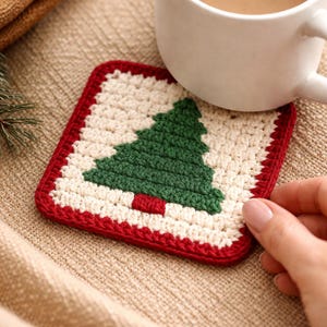 May include: A square crochet coaster featuring a cream-coloured centre with a dark green Christmas tree design in the middle. The tree has a small red trunk at the base. The coaster is bordered by a thick, solid red crocheted edge.