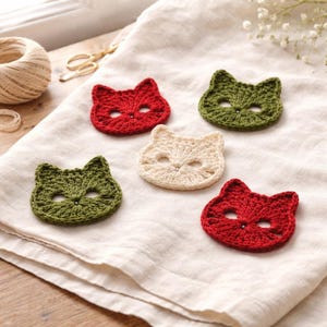 May include: Five crocheted cat face appliques in red, olive green, and cream colors. Each piece features two pointed ears, two eye openings, and a small central stitch detail. The cat-shaped motifs are arranged on a light-colored fabric surface.