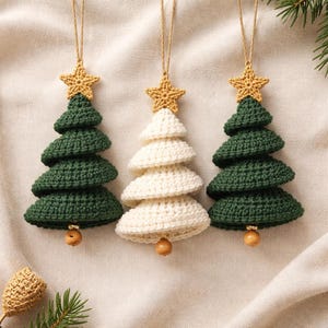Puede incluir: Tres adornos colgantes con forma de árbol de Navidad, dos en verde oscuro y uno en blanco crema. Cada árbol está tejido a ganchillo con capas escalonadas, rematado con una pequeña estrella amarilla y una cuenta de madera colgando.
