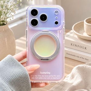 May include: A translucent, iridescent purple smartphone case featuring a circular silver ring stand on the back. The case displays the text "be better" and "luckyday" along with the quote "It is the time you have wasted for your rose that makes your rose so important."