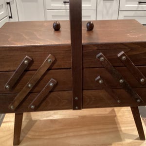 May include: A dark brown wooden sewing box with a cantilevered accordion-style design, featuring a central carrying handle, two small round knobs on the top lid, and four angled legs. The box has visible metal rivets connecting the wooden slats.