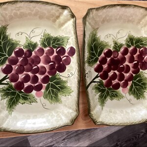 May include: Two rectangular serving platters featuring a painted design of clusters of purple grapes with green leaves and vines on a cream-colored background. The edges of the platters have a textured, scalloped border with a light green wash.