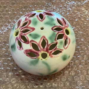 May include: A spherical ceramic flower frog with a white base, featuring hand-painted pink flower motifs and green foliage accents. The surface is punctuated by multiple cut-out petal-shaped openings and small circular holes for stems.