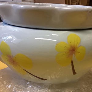 May include: A white ceramic bowl with a wide, flared rim and a rounded body. The exterior surface features a hand-painted design of yellow flowers with brown stems and centers. The bowl has a smooth, glossy finish and a wide opening at the top.