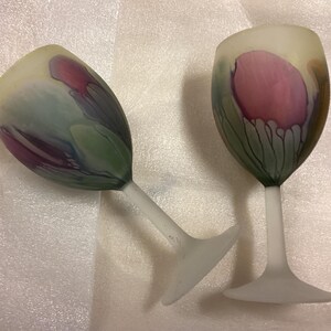 May include: Two frosted glass wine glasses featuring a hand-painted floral design with soft hues of pink, purple, and green. The stems and bases are made of a matte, opaque white glass, while the bowls display a translucent, painterly finish.